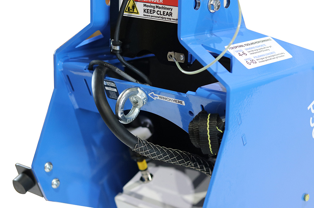 Close-up of a blue industrial machine with a metal winch hook labeled 'WINCH HERE', hoses, and warning labels.